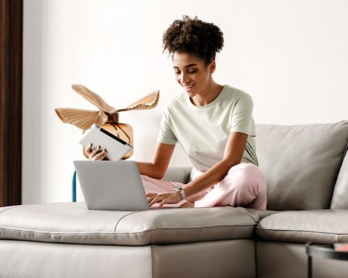 Young black woman using tablet computer while working with laptop